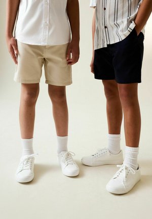 Beeži ja tumesinised lühikesed püksid, mida kannavad kaks inimest, kombineeritud valgete vabaajajalatsite ja soonikuga sokkidega, seisavad kõrvuti neutraalsel taustal.