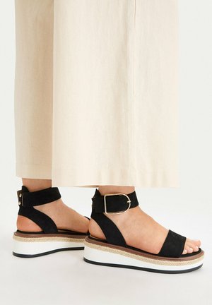 Füße in schwarzen Plateau-Sandalen mit Knöchelriemen und goldenen Schnallen, kombiniert mit cremefarbenen, weit geschnittenen Hosen.