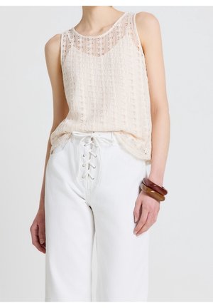 Femme portant un haut en dentelle beige sans manches, rentré dans un pantalon blanc taille haute avec un laçage sur le devant, portant trois larges bracelets en résine au poignet droit.