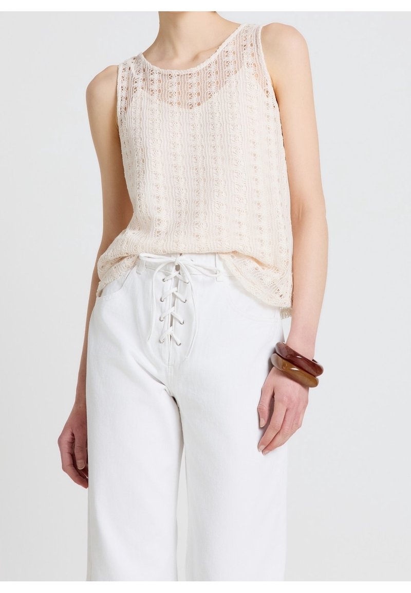 Femme portant un haut en dentelle beige sans manches, rentré dans un pantalon blanc taille haute avec un laçage sur le devant, portant trois larges bracelets en résine au poignet droit.