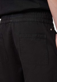 Shorts en tissu noir avec un waistband élastique, deux poches arrière et des coutures ton sur ton. Le tissu a une texture lisse et un fini mat.
