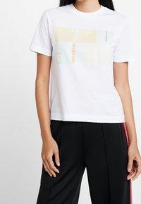 A person wears a white t-shirt featuring a multi-colored Calvin Klein logo graphic, paired with black pants with red stripes.