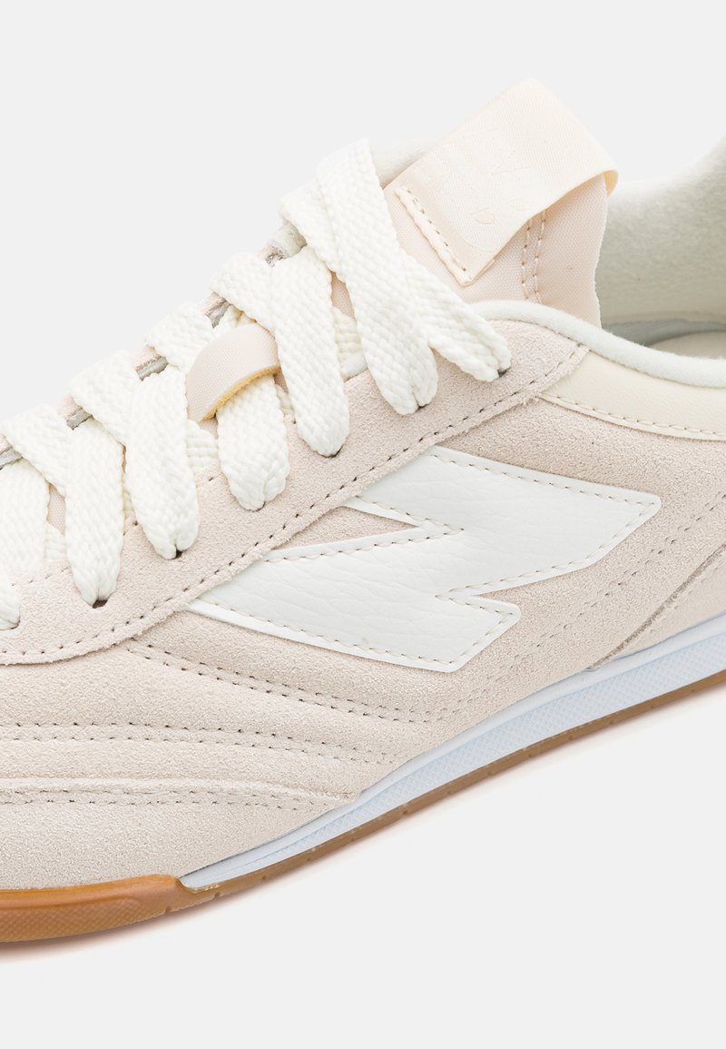 Sneaker beige clair avec une tige en daim, logo en cuir blanc et semelle en caoutchouc texturée. Fermeture à lacets avec des lacets plats blancs et un col rembourré.