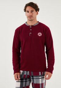 Burgunderfarbener Longsleeve mit drei Knöpfen, grauem Kragen und Brustlogo; kombiniert mit karierten Schlafanzughosen in Rot, Grau und Weiß.