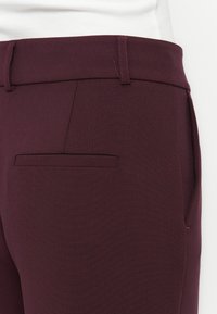 Burgundské kalhoty s hladkým povrchem, přiléhavým střihem, plochou přední kapsou a minimalistickým pasem. Žádné viditelné kování nebo ozdoby.