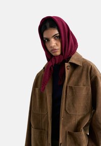 Femme portant un foulard en tricot bordeaux noué sous le menton et une veste en daim marron avec de grandes poches devant sur un fond clair.