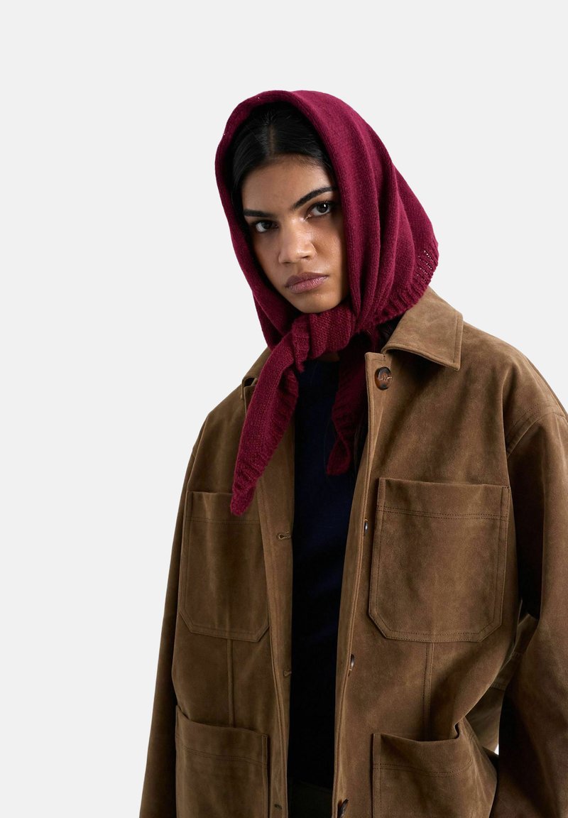 Femme portant un foulard en tricot bordeaux noué sous le menton et une veste en daim marron avec de grandes poches devant sur un fond clair.