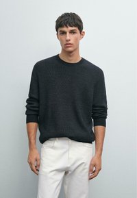 Junger Mann in einem dunkelgrauen Strickpullover und weißen Hosen, der vor einem einfarbigen hellgrauen Hintergrund steht.