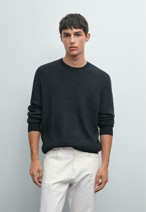 Young man wearing a dark gray knit sweater and white pants standing against a plain light gray background.