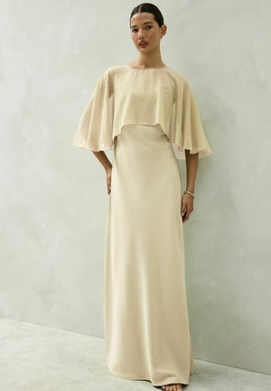 Frau trägt ein langes, beiges, fließendes Kleid mit einem lockeren, durchsichtigen Cape und steht vor einer schlichten hellgrauen Wand.