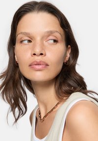 Kobberforbindelses kædetrøje med glat tekstur, båret over en lysebeige ærmeløs top. Modellen har mellemlangt brunt hår og naturlig makeup.