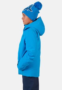 Jasně modrá voděodolná bunda s kapucí, s hladkým povrchem. Spárována s modrou pletenou čepicí zdobenou bambulí a grafickým vzorem.