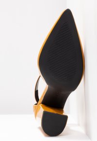 Chaussure à talon haut en cuir verni jaune avec un bout pointu, dotée d'une semelle extérieure en caoutchouc noir avec des rainures de traction et d'un talon ouvert.