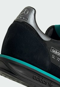 Black suede sneaker with silver accents, turquoise sole, and 'adidas SL '72' label. Features a smooth texture and a low-profile design.