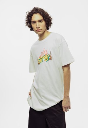 Camiseta blanca de gran tamaño con un gráfico colorido de "GRIMY" y acentos de llamas. Tiene mangas cortas y un corte holgado. Se lleva con pantalones cortos oscuros.