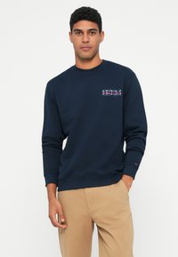 Mlad moški v temno modrem puloverju z napisom "1985" in bež hlačami, stoji ob belem ozadju z eno roko v žepu.