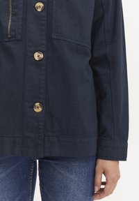 Mørkeblå jakke laget av teksturert stoff, med store bronseknapper, en frontglidelås og sidelommer. Ermene er lett innsvingt.