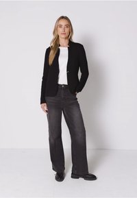 Zwarte blazer over een witte blouse, gecombineerd met donkergrijze wijde denim en zwarte laarzen. Glad stofoppervlak met minimalistische ontwerpelementen.