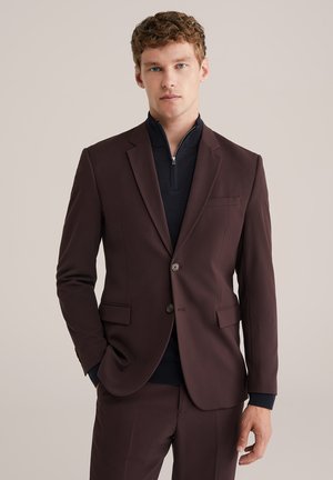 Young man wearing a dark brown suit with a navy zip-neck sweater, standing against a plain beige background.