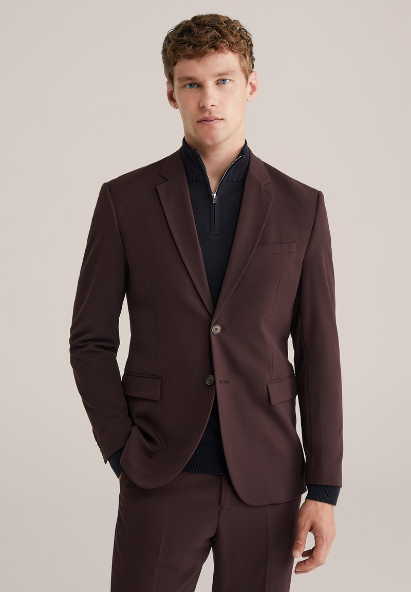 Hombre joven vestido con un traje marrón oscuro y un suéter de cuello zip azul marino, de pie frente a un fondo beige liso.