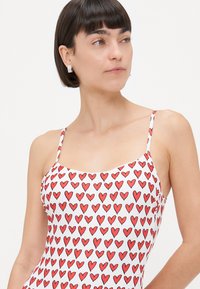 Fehér fürdőruha ismétlődő piros szív mintával, vékony pántokkal és kerek nyakkivágással. Sima anyag, mely testhezálló kialakítású.