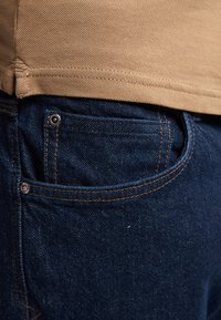Jeans de mezclilla en azul oscuro con costuras naranjas contrastantes, que cuentan con bolsillos laterales y un bolsillo para monedas. La camisa es de un tejido texturado en color beige en la cintura.
