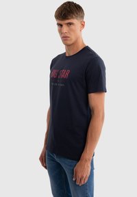 Man draagt een marineblauw T-shirt met de tekst "BIG STAR" en een blauwe spijkerbroek, staand zijwaarts tegen een lichtgrijze achtergrond.