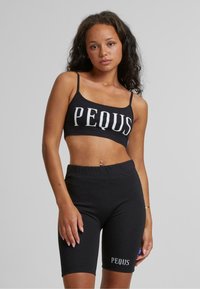 Svart croppet topp og matchende høytalede shorts laget av elastisk bomull, med en stor hvit "PEQUS"-logo på begge plagg.