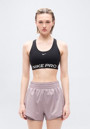 Fiatal nő rövid, nedves hajjal, fekete Nike Pro sportmelltartót és mályva színű sportnadrágot viselve, egyszínű háttér előtt áll.