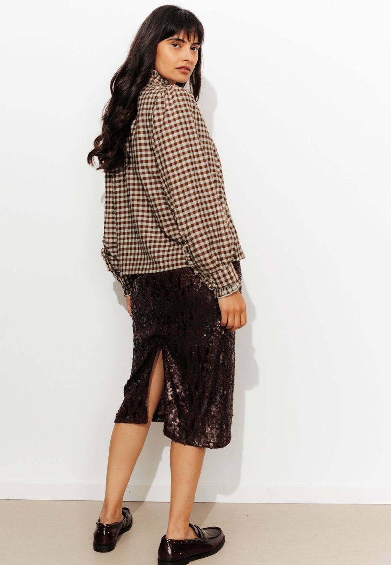 Femme aux cheveux longs et foncés, portant une blouse à carreaux marron, une jupe noire en sequins avec une fente à l'arrière, et des mocassins foncés, se tenant contre un mur blanc.