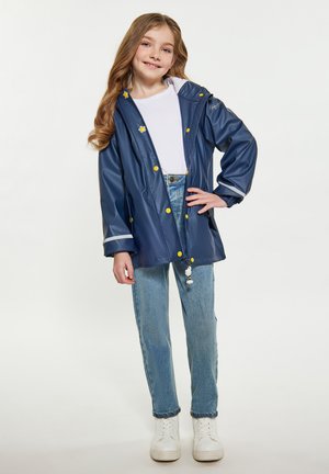 Giacca impermeabile della marina con bottoni gialli, cappuccio con cordino e dettagli riflettenti, abbinata a jeans azzurri e sneaker bianche.