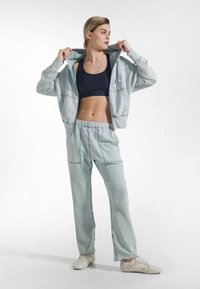 Chándal de tela suave de color azul claro con una sudadera con capucha con cremallera y pantalones con cintura elástica, combinado con un sujetador deportivo oscuro y zapatillas claras.