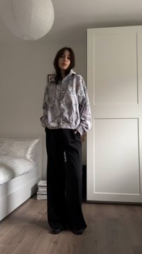 Γκρι φουσκωτό φούτερ με φερμουάρ και χαλαρή εφαρμογή, συνδυασμένο με μαύρο παντελόνι φαρδιάς γραμμής. Το δωμάτιο διαθέτει λευκό κρεβάτι και ντουλάπα.