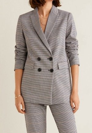 Frau trägt ein grau kariertes, zweireihiges Blazer mit schwarzen Knöpfen und passenden Hosen, stehend vor einem neutralen Hintergrund.