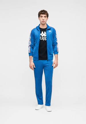 Mladý muž stojaci oblečený v modrej šuštiakovej súprave značky Kappa, čiernom tričku s logom a bielych teniskách, na jednoduchom pozadí.