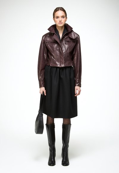 Jeune femme portant une veste en cuir brun foncé, une jupe noire longueur genoux, des bottes noires montantes, tenant un sac à main noir, debout avec une expression neutre.