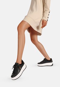 Sneakers in pelle nera con suola piatta bianca, dotati di lacci e dettagli in metallo, abbinati a un vestito oversize beige.