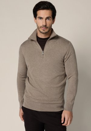 Mężczyzna o krótkich ciemnych włosach, ubrany w beżowy sweter rozpinany na zamek, nałożony na czarną koszulkę, stojący na jednolitym jasnym tle.