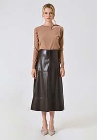 Braunes Langarmoberteil und einen hoch-taillierten braunen Lederrock mit ausgestelltem Saum, der über Nahtdetails verfügt. Schuhe mit spitzer Form in neutralen Farbtönen rundet das Outfit ab.
