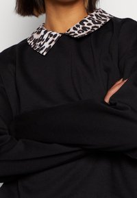 Persona con los brazos cruzados que lleva una camiseta de manga larga negra con un cuello de estampado de leopardo en beige y negro.