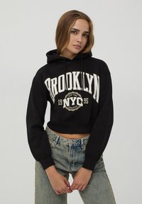 Felpa corta nera con testo bianco "BROOKLYN NYC 1995"; materiale in cotone; presenta una tasca frontale e cappuccio con cordini; vestibilità rilassata.