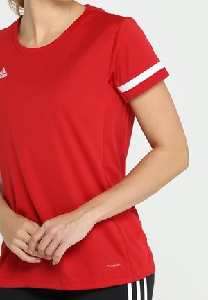 Vrouw draagt een rood sportshirt met korte mouwen en een witte streep op de mouw, en een zwarte broek met witte strepen, staand met de handen gevouwen.