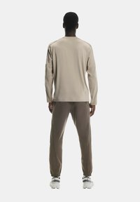 Cămașă bej cu mânecă lungă și pantaloni jogger maro dintr-un material moale. Pantalonii au manșete elastice și buzunare. Adidași albi și gri completează ținuta.