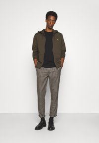Felpa con zip verde oliva, tasche a marsupio, t-shirt blu scuro sotto, pantaloni a quadri grigi, mocassini neri e maniche arrotolate.