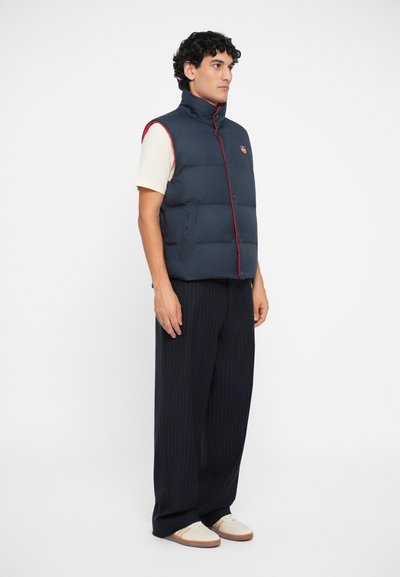 Navy puffer vest with red stitching and high collar, featuring button closure and side pockets, worn over a cream T-shirt and pinstripe pants.
