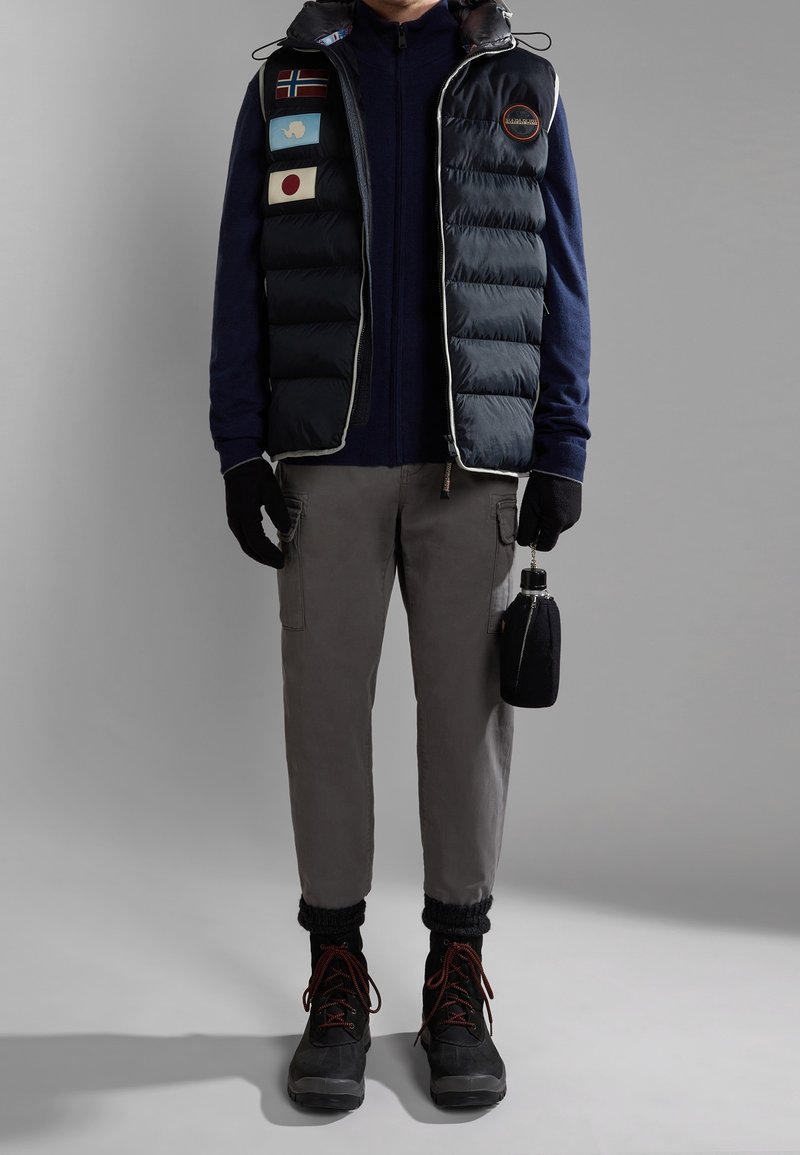 Personne portant un gilet matelassé bleu marine avec des écussons, une chemise à manches longues bleu marine, un pantalon cargo gris, des gants noirs et des bottes noires tenant une gourde noire.