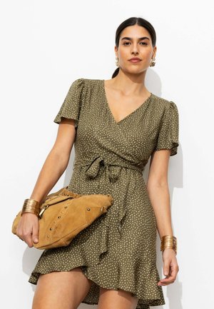 Femme portant une robe portefeuille olive à pois, tenant une pochette en daim marron cloutée, avec des bracelets et des boucles d’oreilles dorées, debout devant un fond blanc.