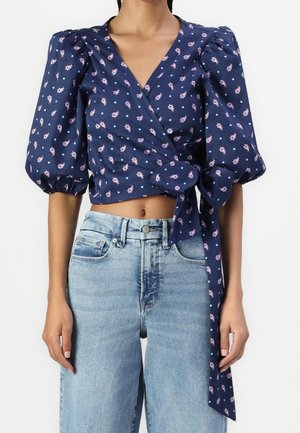 Woman wearing a navy blue wrap crop top with pink floral and white polka dots, puff sleeves, tied at the waist, paired with light blue jeans.