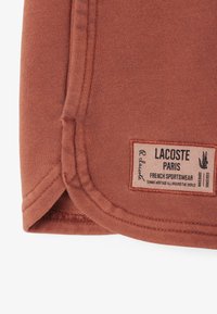 Tissu couleur rouille avec une texture lisse et un ourlet cousu. Présente une étiquette avec "Lacoste Paris" et un logo de crocodile.