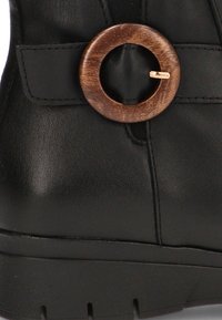 Botín negro de cuero hasta el tobillo con un acento circular de madera y paneles laterales elásticos para facilitar su uso. Superficie texturizada y suela resistente.
